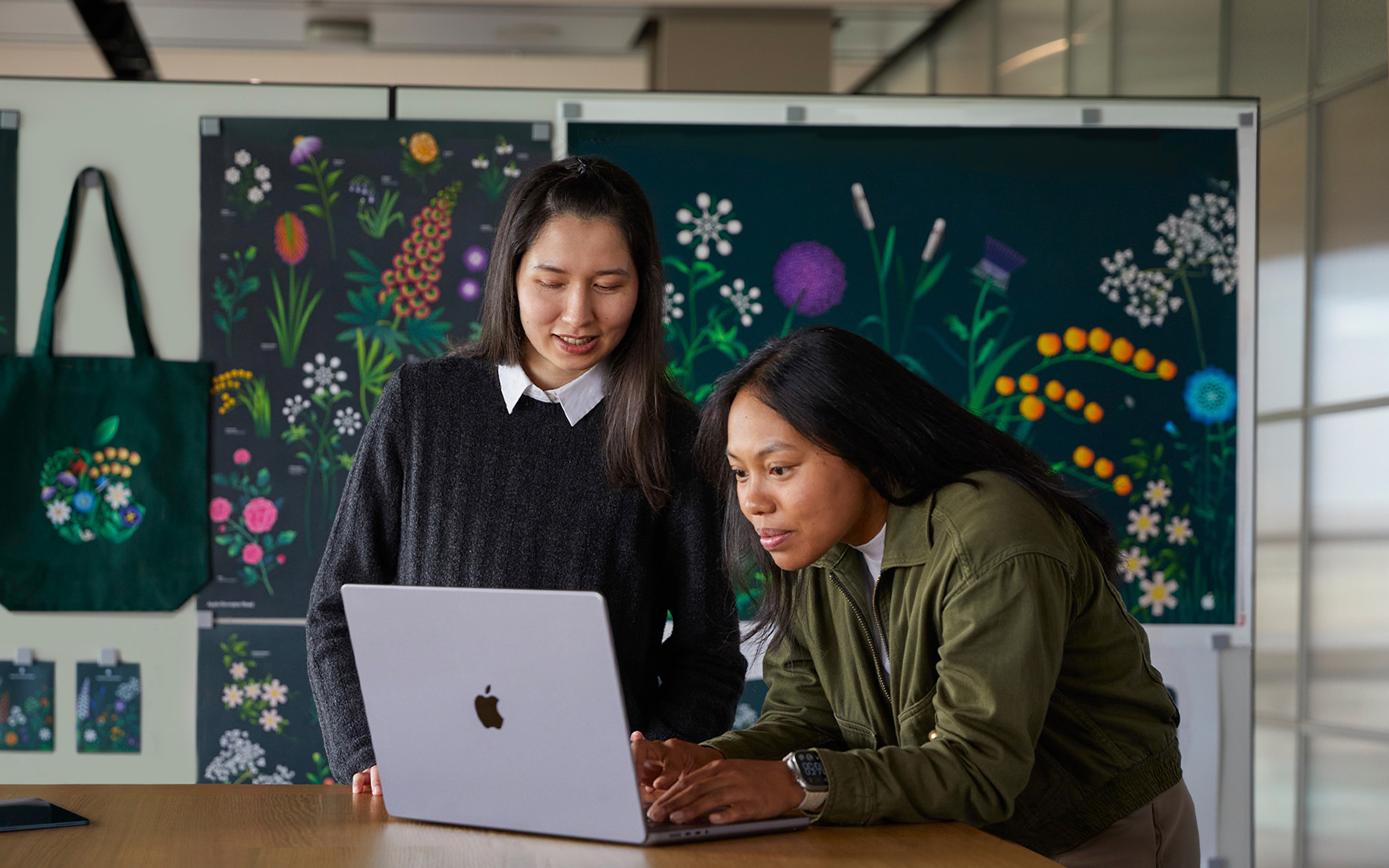 Dos personas del equipo de Marketing de Apple trabajan juntas en una MacBook delante de carteles diseñados por su equipo.