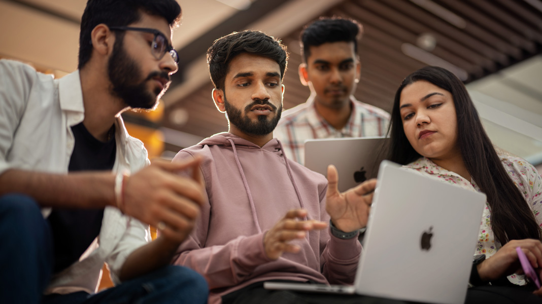 Empat orang mahasiswa tampak berkumpul mengelilingi sebuah MacBook sambil berdiskusi satu sama lain.