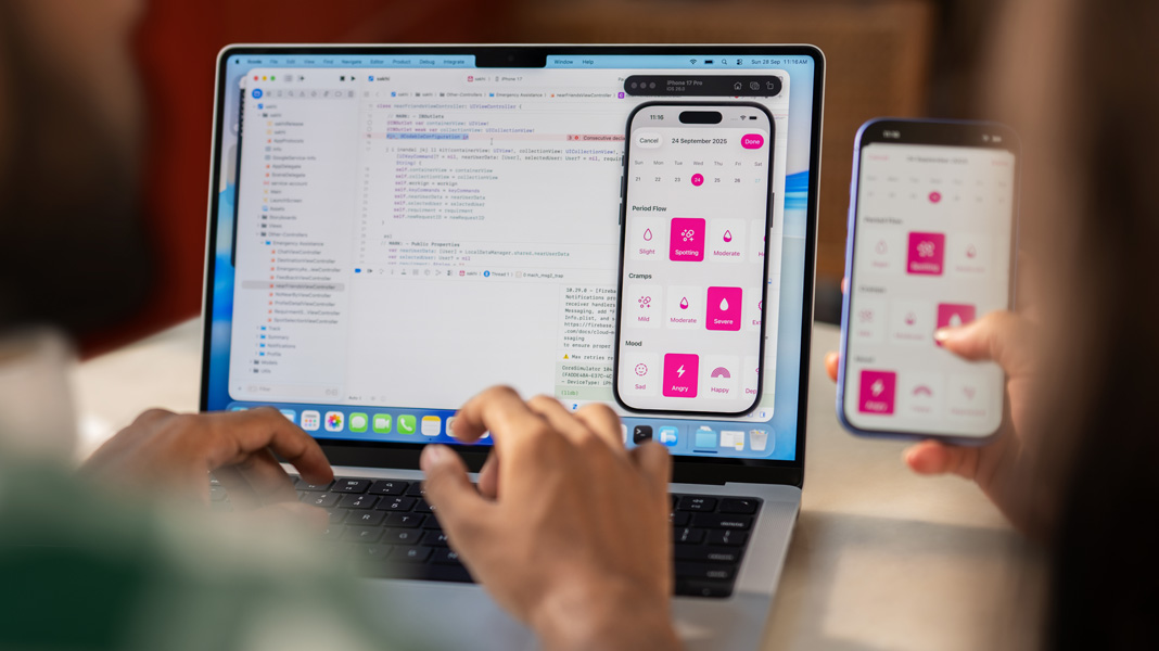 Tampilan dari balik bahu seseorang yang sedang mengetik di MacBook yang menjalankan Xcode, sementara seseorang memegang iPhone yang menampilkan aplikasi Sakhi, yang juga ditampilkan di MacBook.