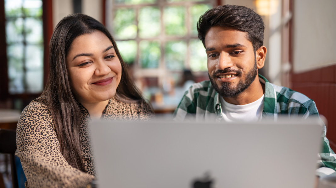 Dua mahasiswa tersenyum hangat sambil melihat MacBook yang terbuka di ruangan yang cerah dan berudara segar.
