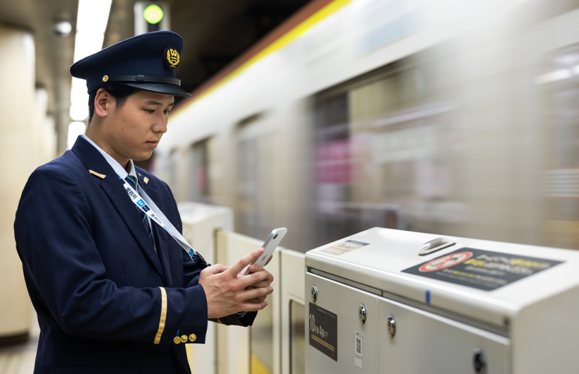 En stationsvärd står på plattformen och använder sin iPhone för att läsa av en qr-kod medan ett tåg kör förbi.