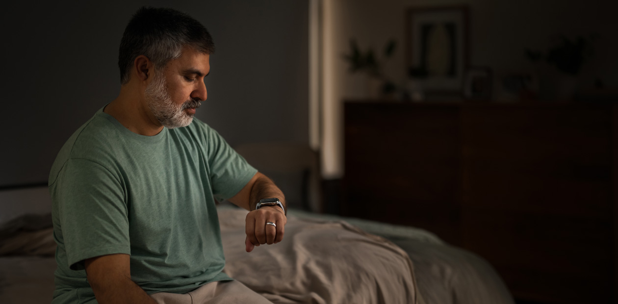 رجل يجلس في السرير يتفقد ساعته Apple&nbsp;Watch.‏