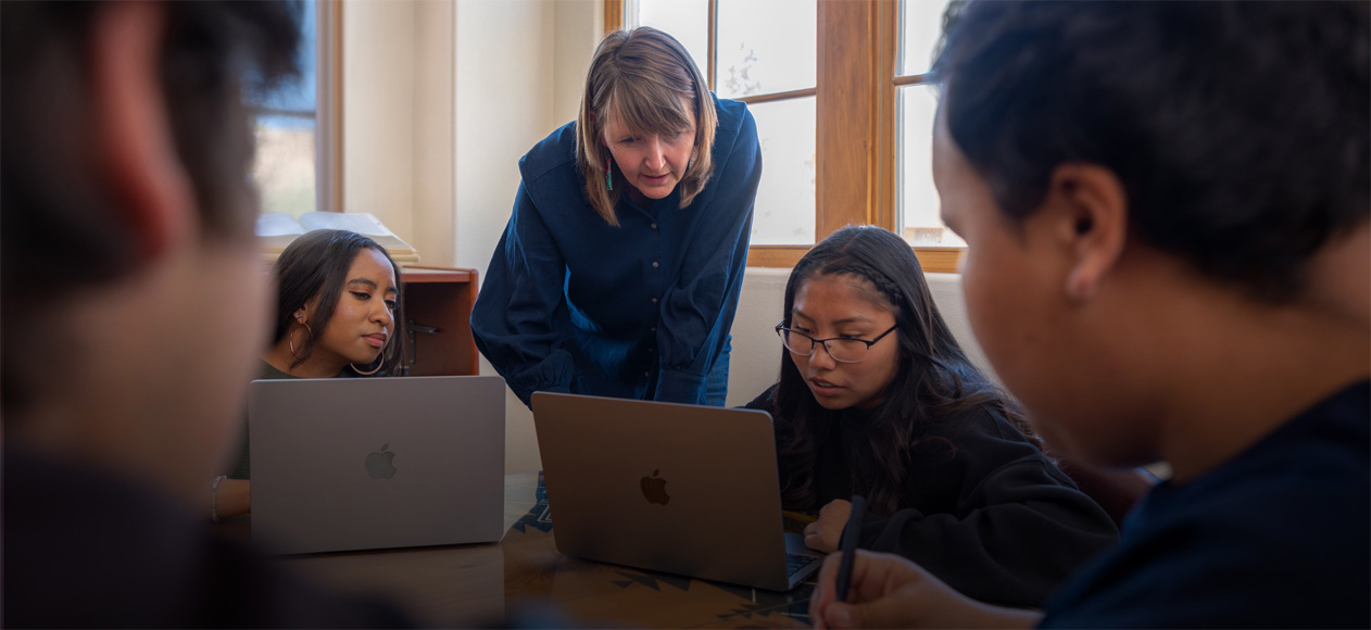 Teacher leaning over to help students working on MacBooks.